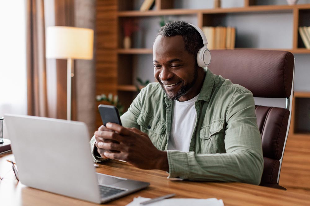 Happy middle aged african american man in casual and wireless headphones typing on smartphone with laptop